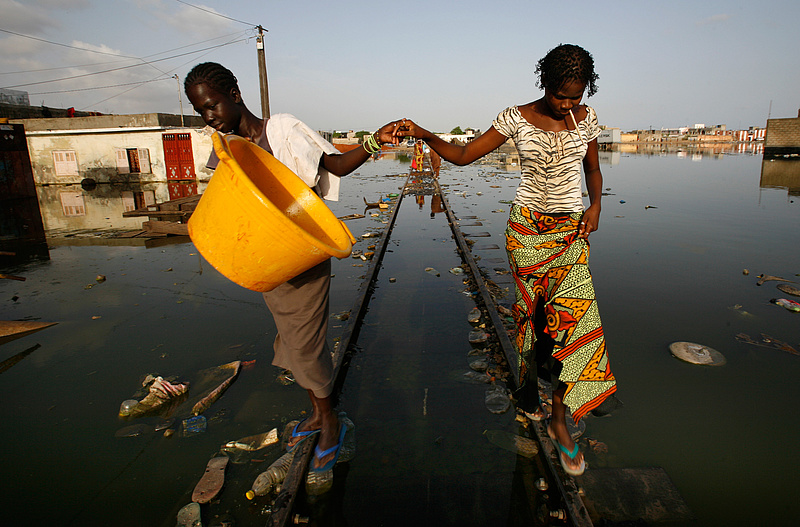 Zwei Frauen balancieren nebeneinander auf einem von Wasser umfluteten Bahngleis. Im Wasser schwimmt Plastikmüll.
